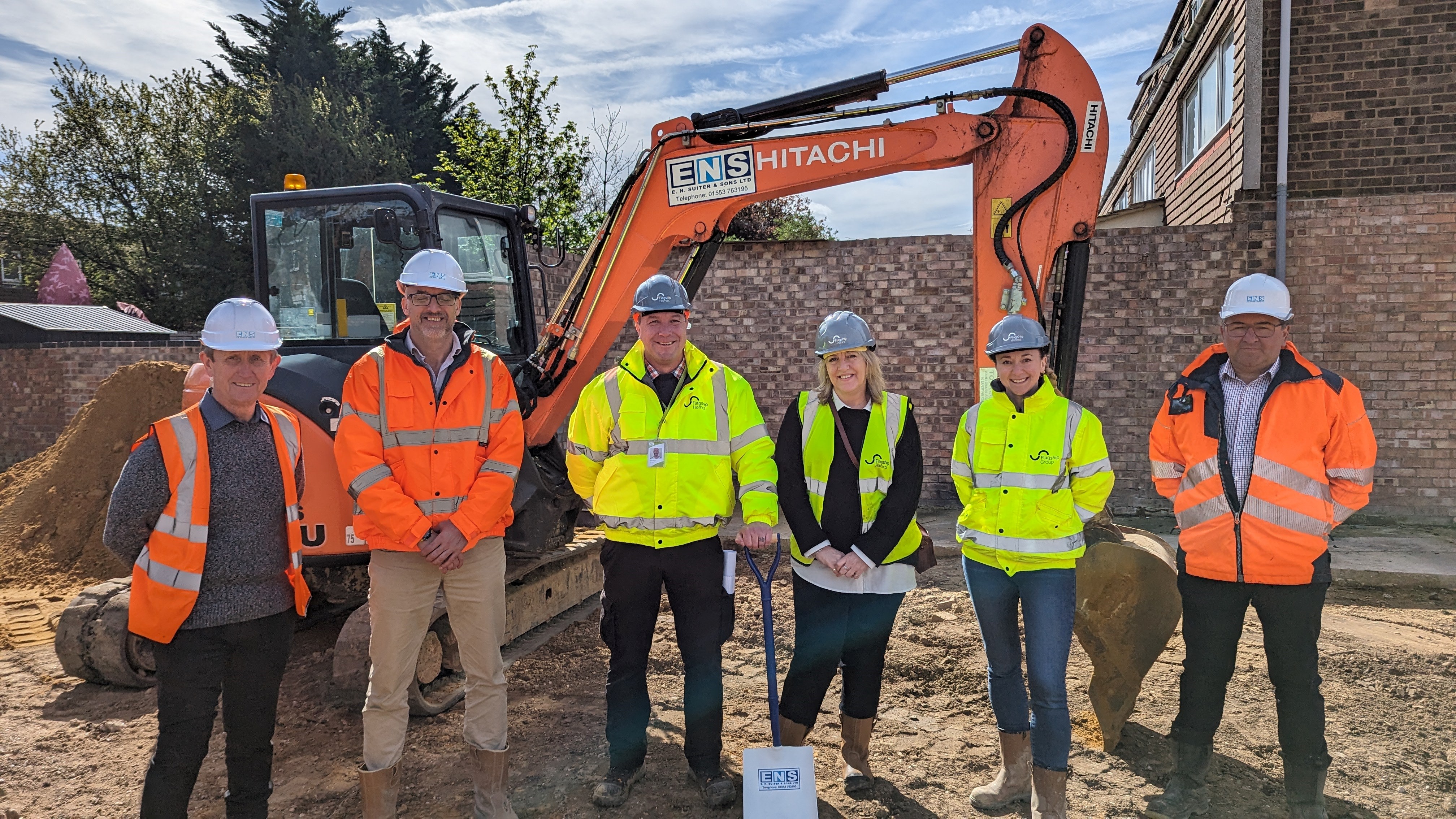 L-R: Martin Kearns, Steven Gould (EN Suiter), Simon Floor, Lisa Davis. Anna Cook-Bacon (Flagship Homes),  and Ian Reeve (En Suiter) on one of the garage sites to be turned into new affordable homes in Mildenhall. All 6 are standing in hi-vis jackets in front of a small digger
